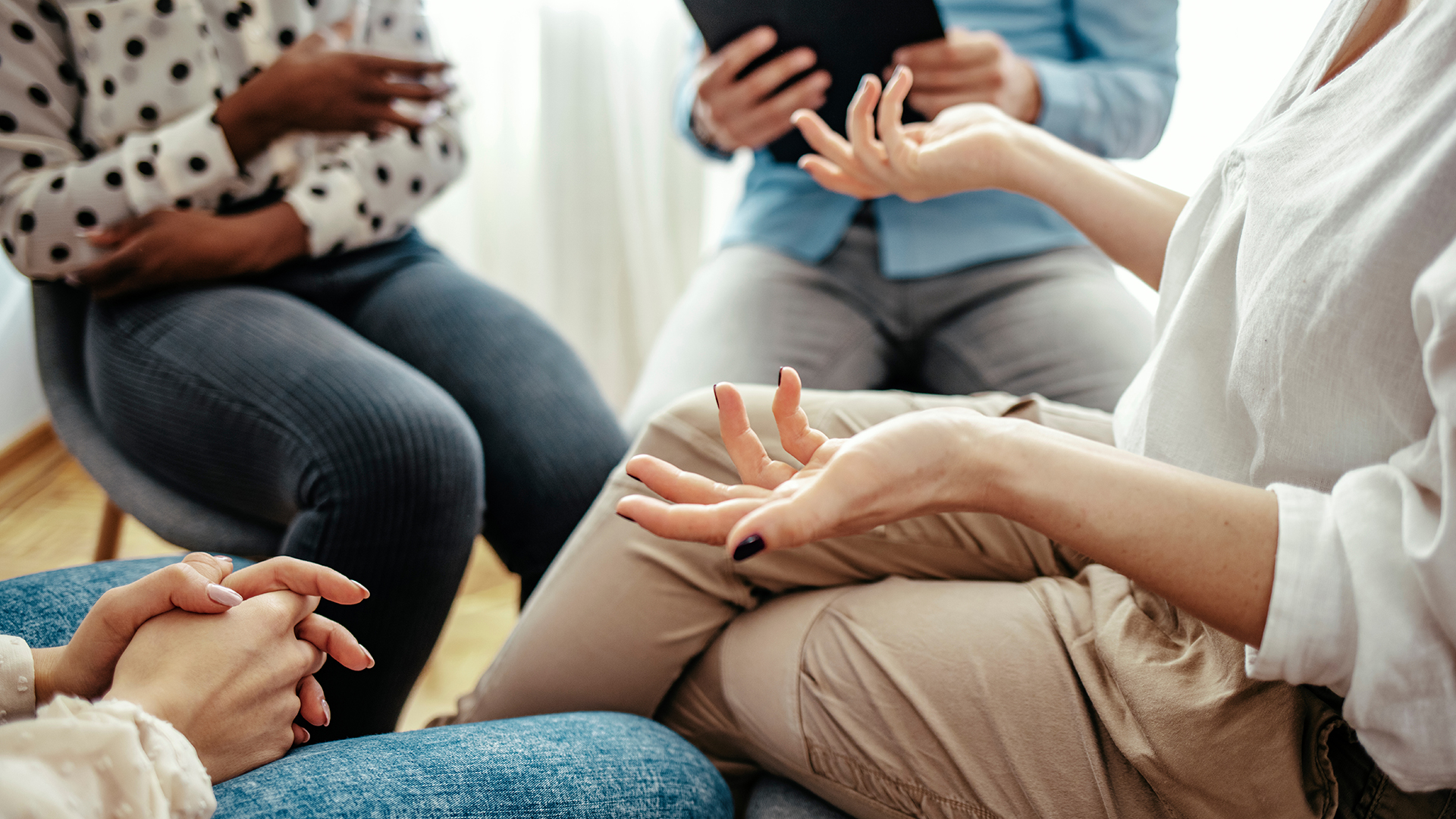  Unrecognizable vulnerable woman participates in group therapy session. Unrecognizable Caucasian person gestures while speaking. Group of people sitting in a circle during therapy. 