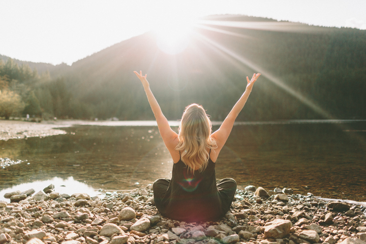 A woman sitting beside a lake with her arms up high.